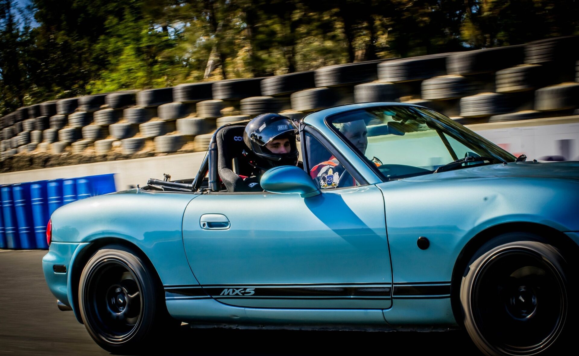Light blue Mazda MX-5 with a driver and passenger wearing helmets on a racetrack with tires in background.