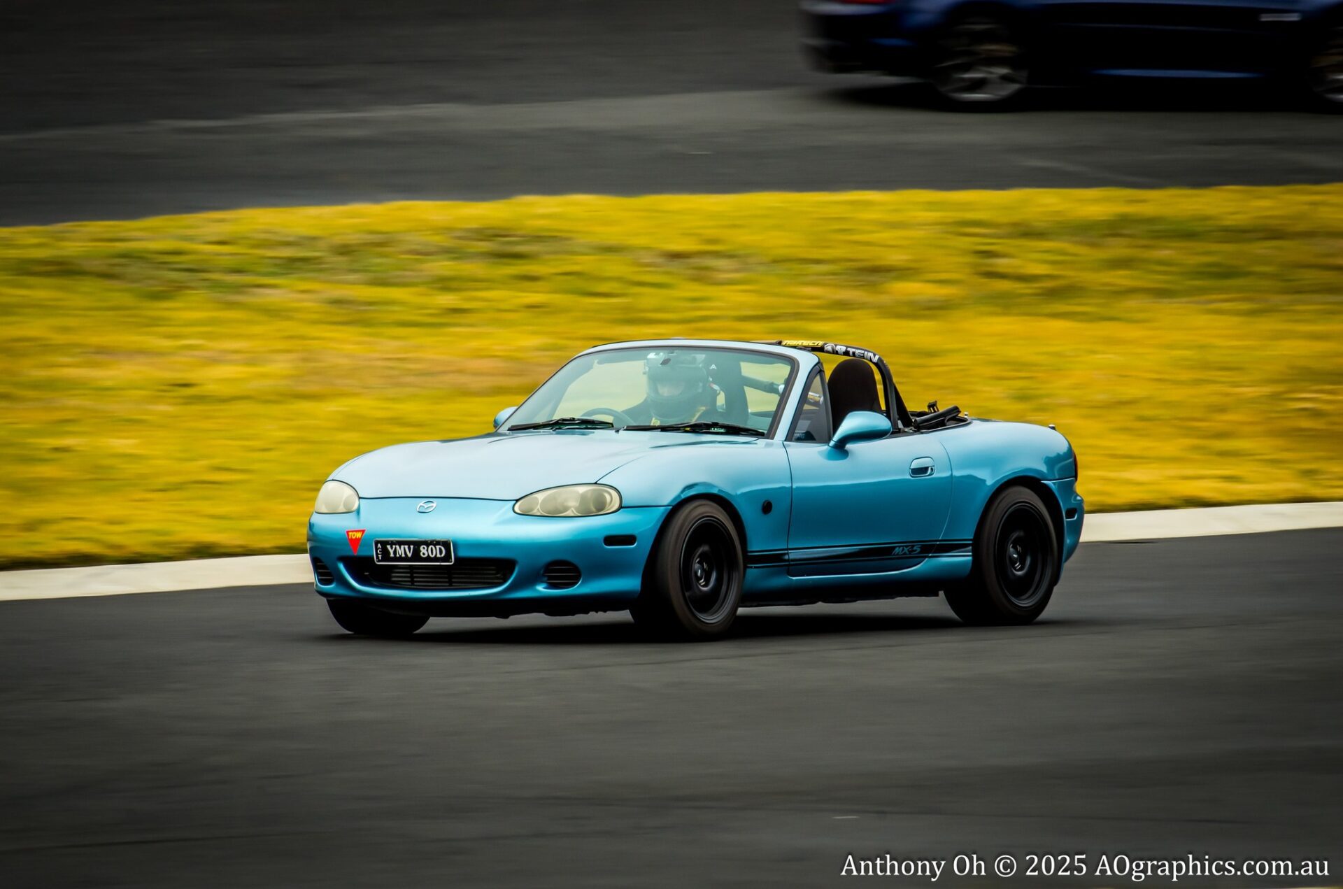 Blue Mazda MX-5 convertible race car with driver wearing a helmet on a racetrack.