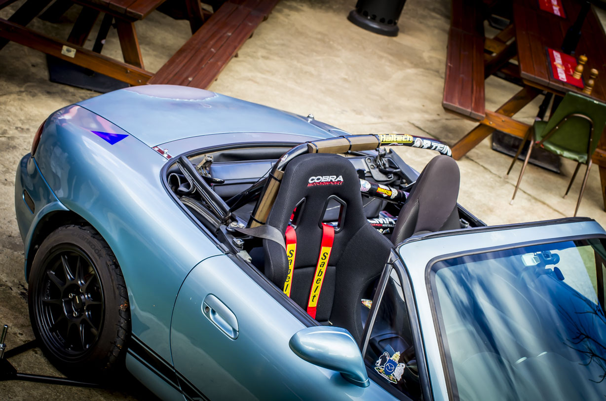 Light blue convertible sports car with racing bucket seat and roll cage, parked near wooden benches.