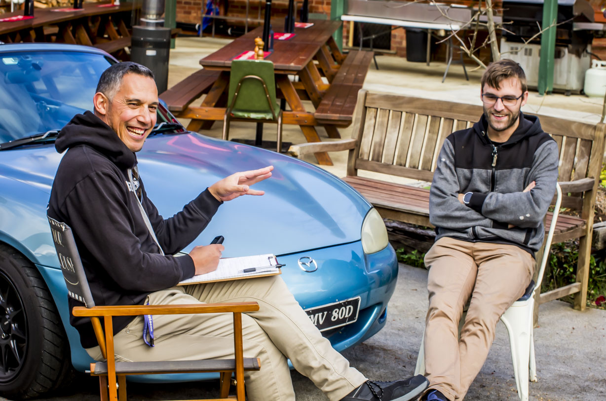 Two men sit outdoors chatting, one holding a clipboard, near a blue car and wooden benches.