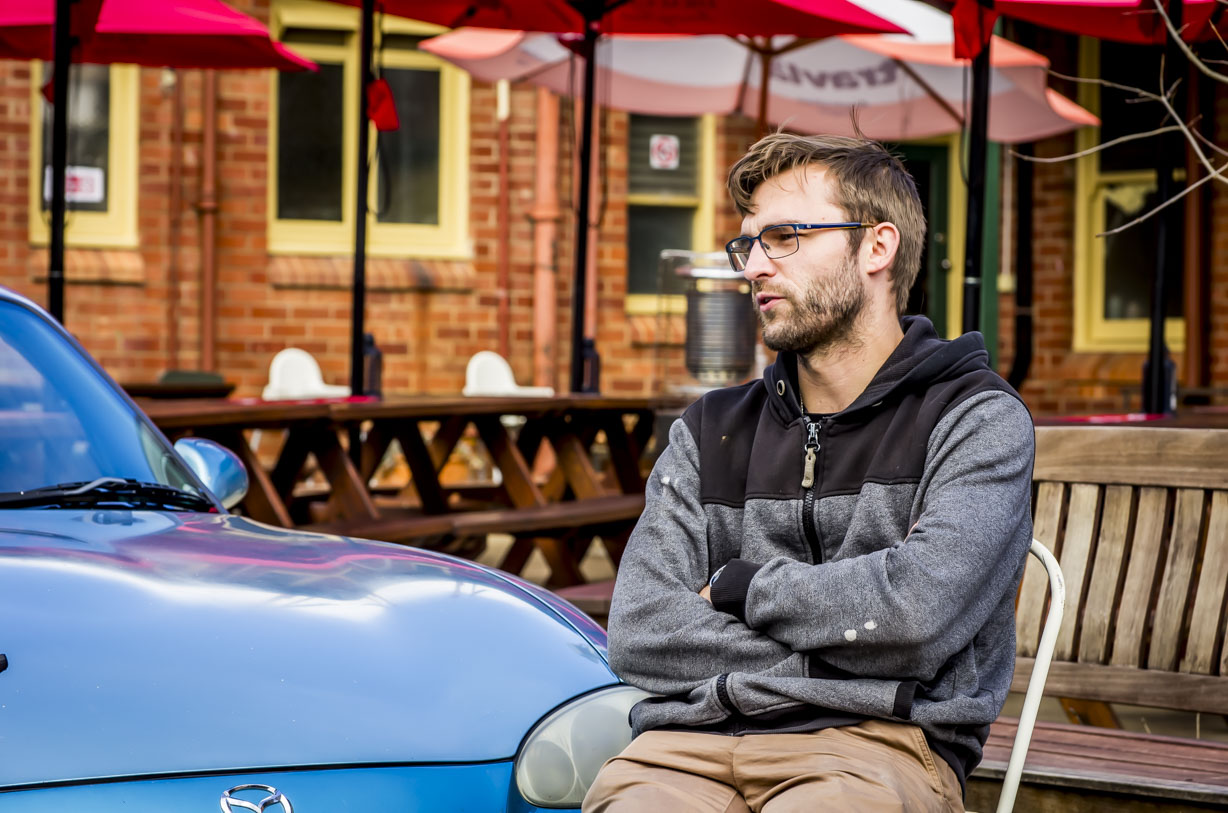 Man wearing glasses and a hoodie sitting on a chair outdoors near a blue Mazda car.