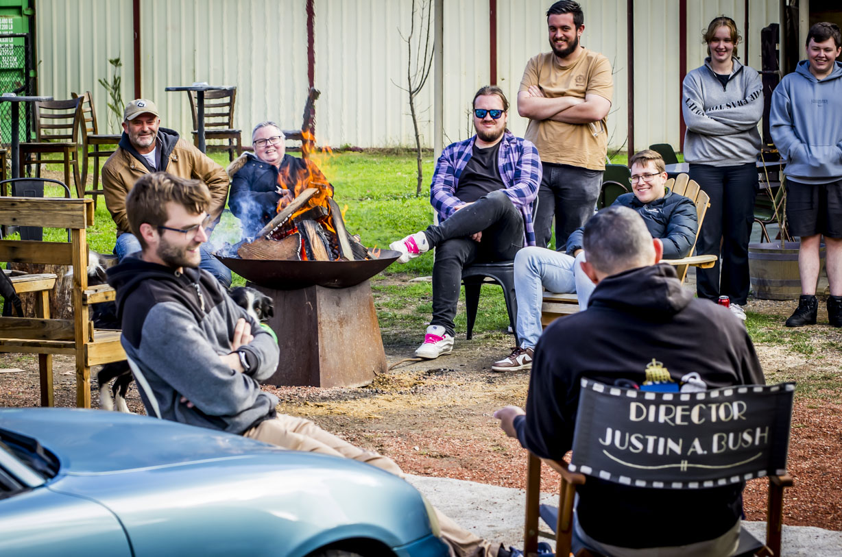 Group of people sitting and standing around a fire pit, socializing outdoors on a sunny day.