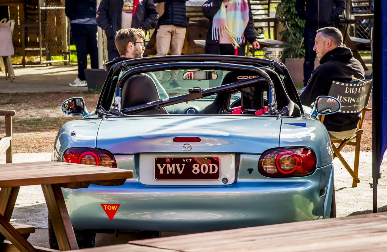 Rear view of a blue convertible Mazda with two men nearby, one sitting on a director's chair.