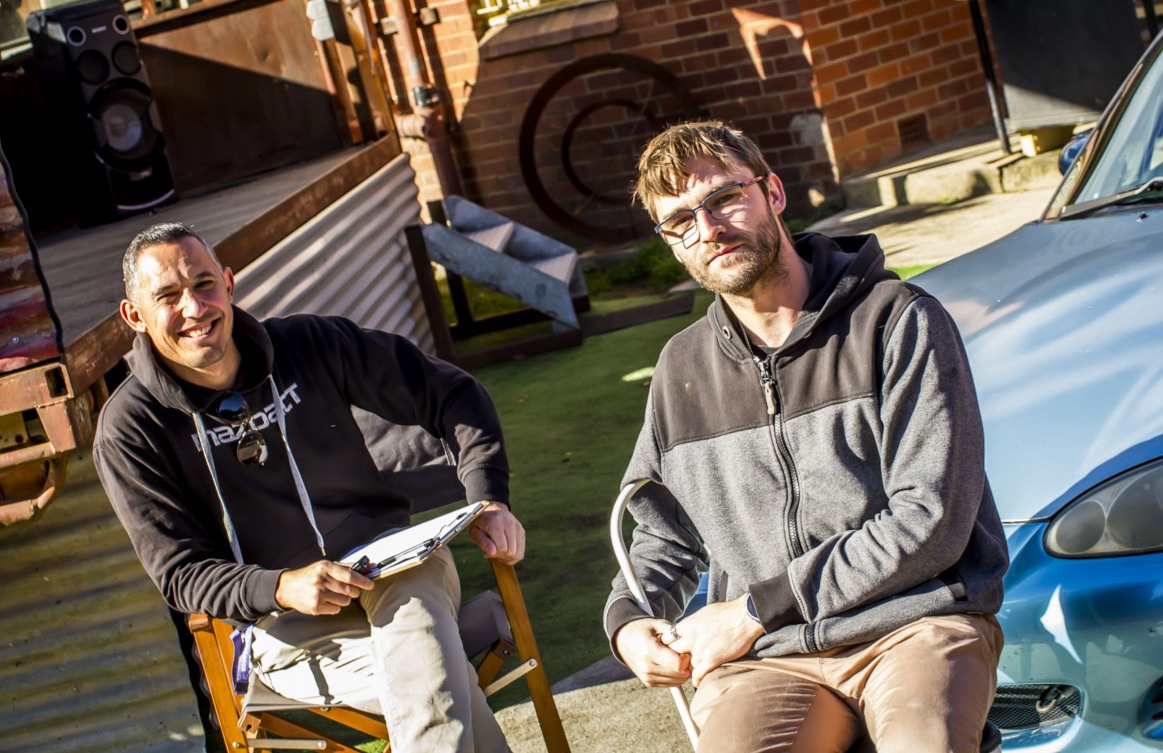 Two men sitting outdoors by a blue car, one holding a clipboard, both in hoodies.