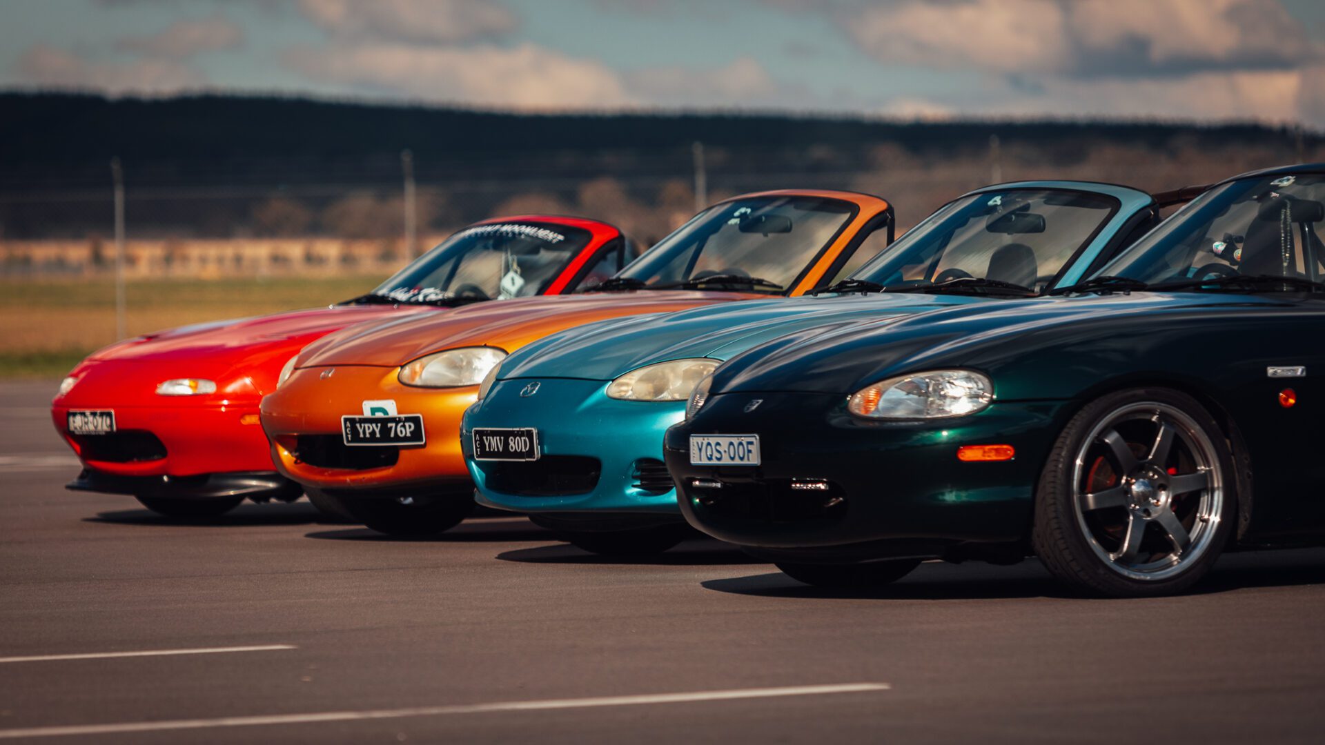 Four colorful Mazda MX-5 convertible cars in red, orange, turquoise, and dark green parked side by side.