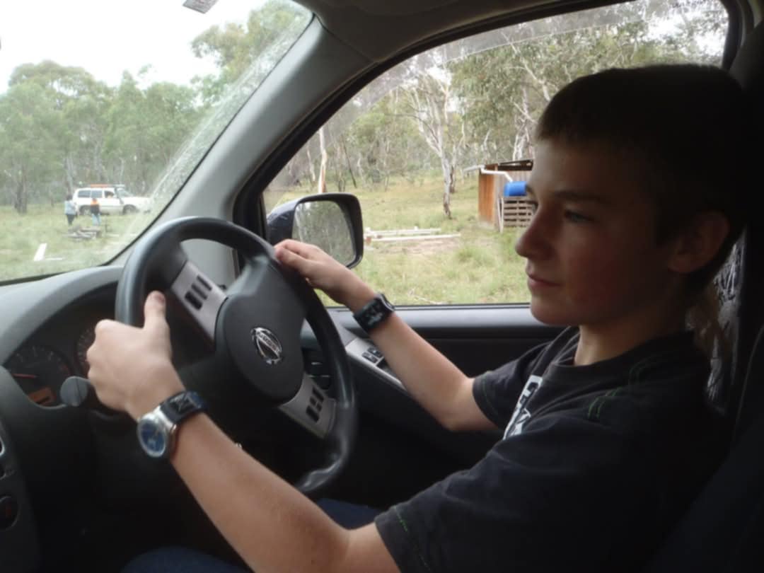 Teen boy sitting in the driver's seat of a car, holding the steering wheel, outdoors.