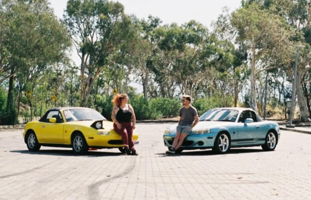 Two people sitting on the hoods of a yellow and a blue convertible cars in a park area with trees.