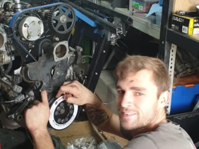 Man working on a car engine part in a workshop with tools and shelves in the background.