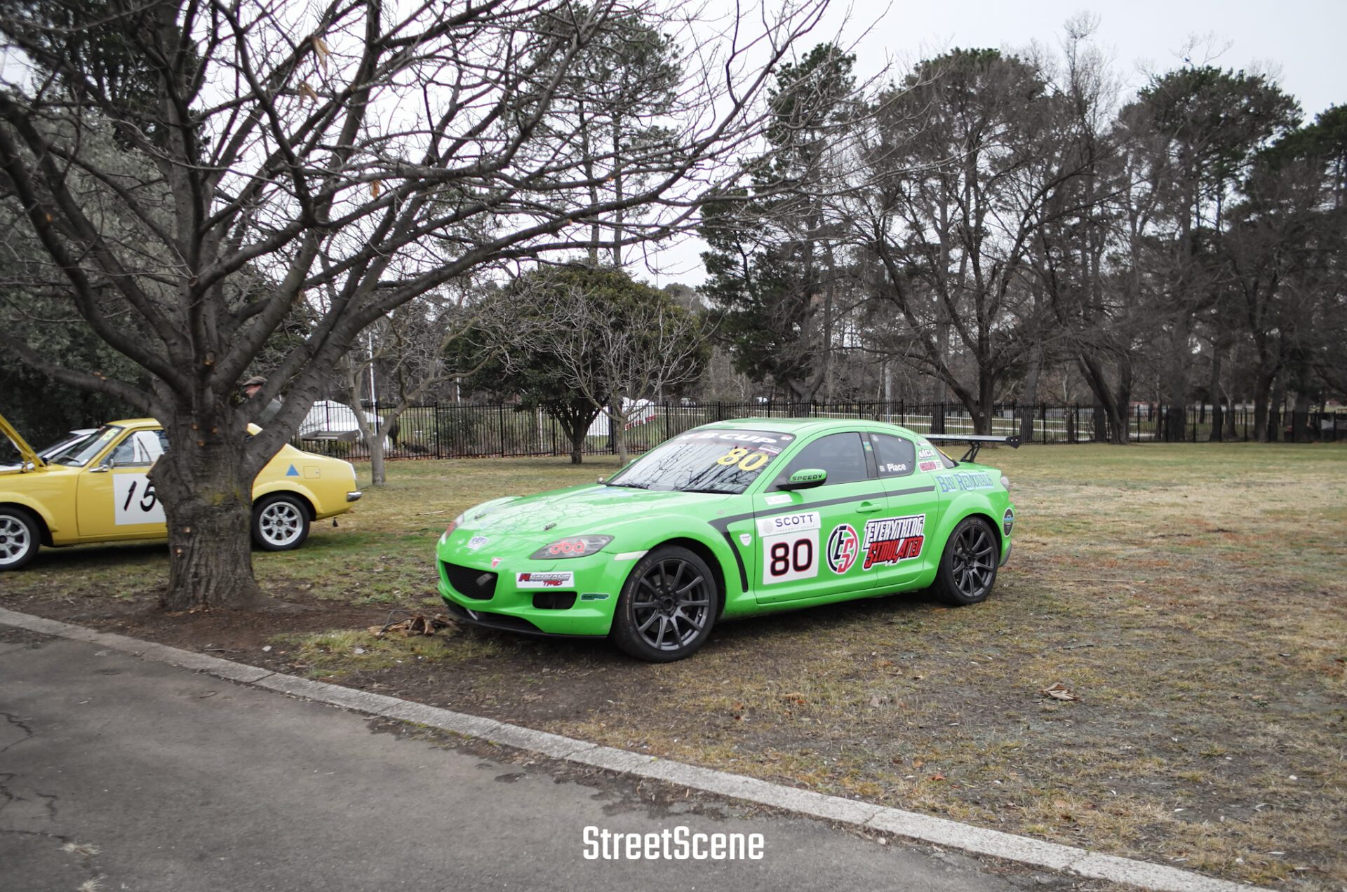 Bright green race car number 80 parked on grass next to a yellow vintage car and leafless trees.