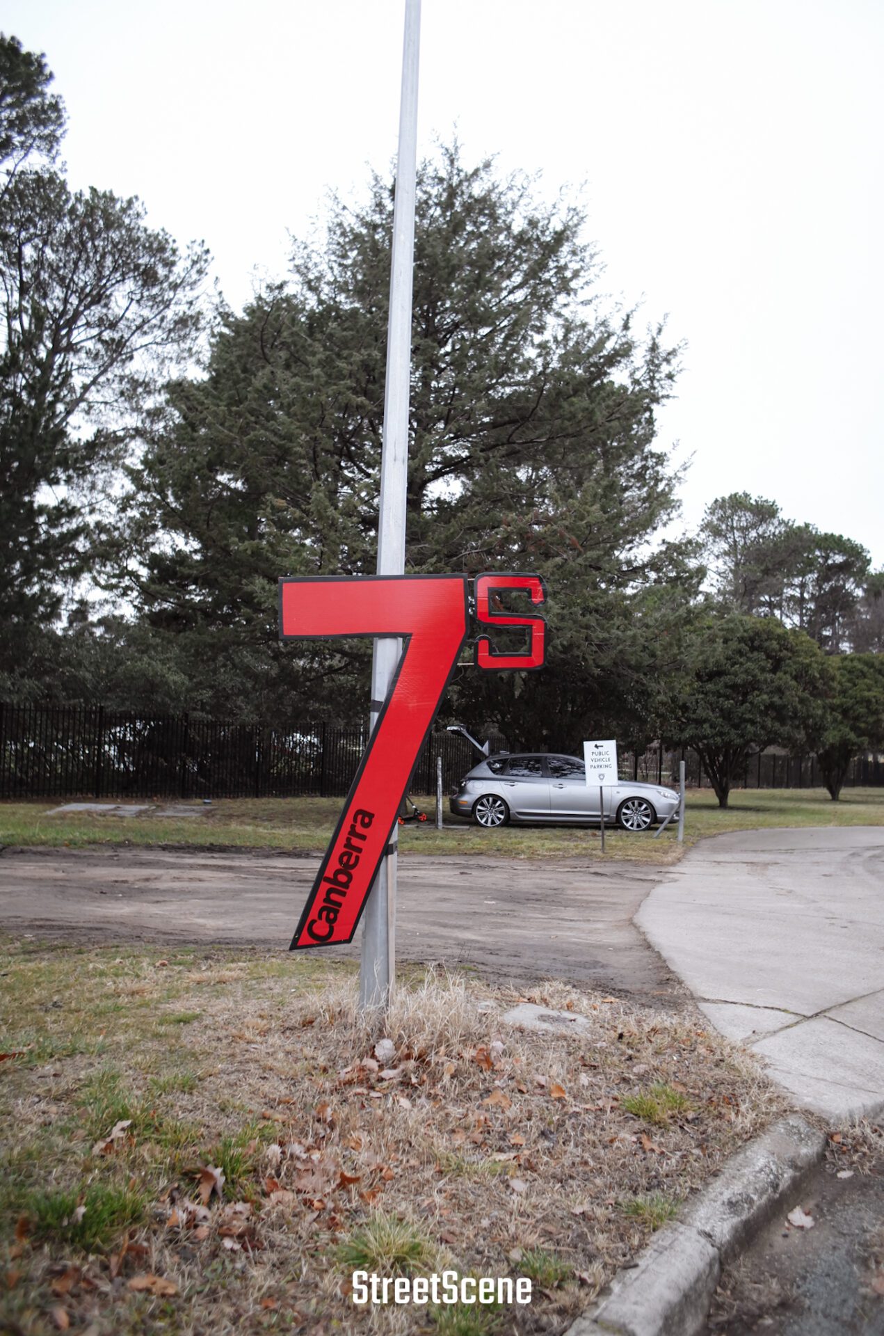 Red "7s Canberra" sign on a pole near a grassy roadside with trees and a parked silver car.