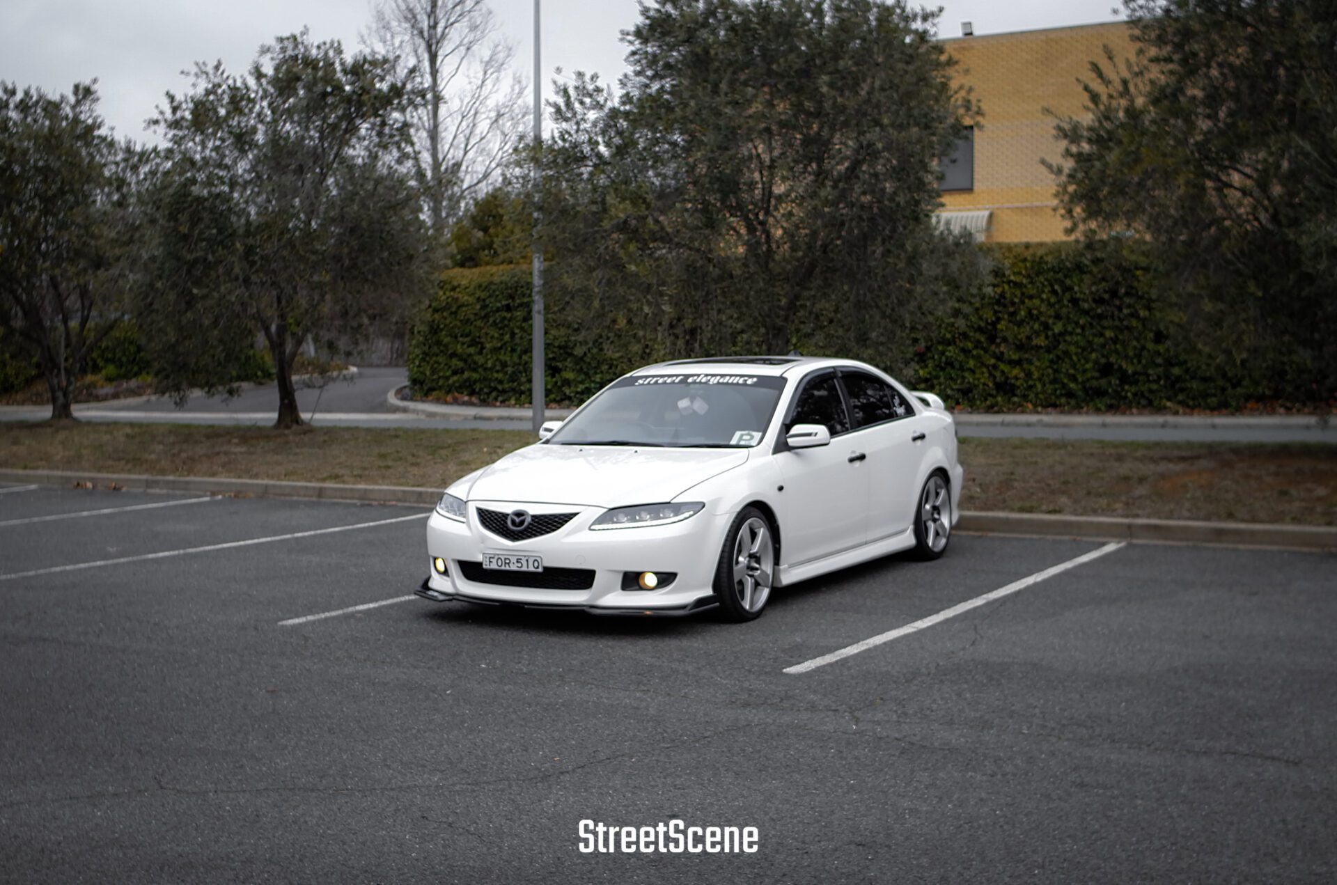 White Mazda sedan with lowered suspension parked in an empty outdoor parking lot.