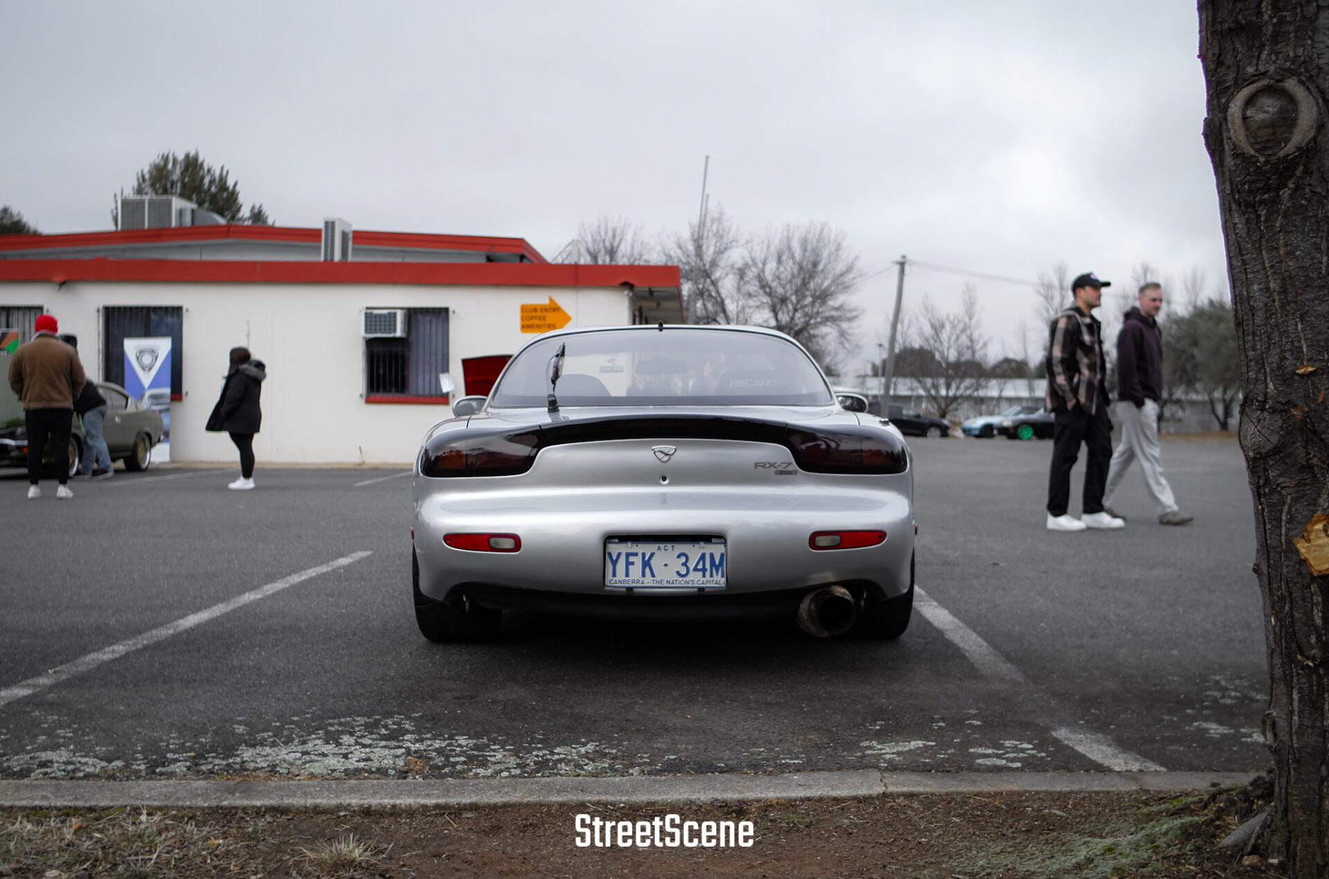 Silver Mazda RX-7 parked in a lot on a cloudy day with people and a building in the background.