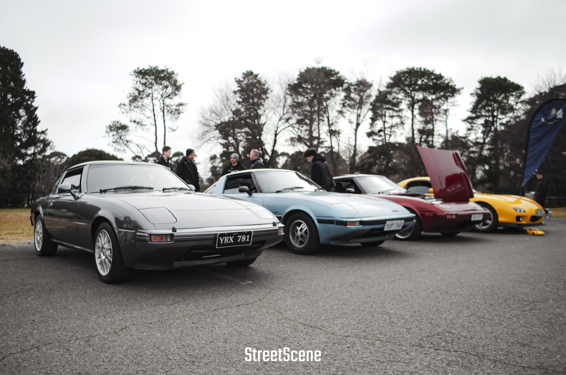 Four vintage sports cars parked in a row with people chatting in the background on a cloudy day.