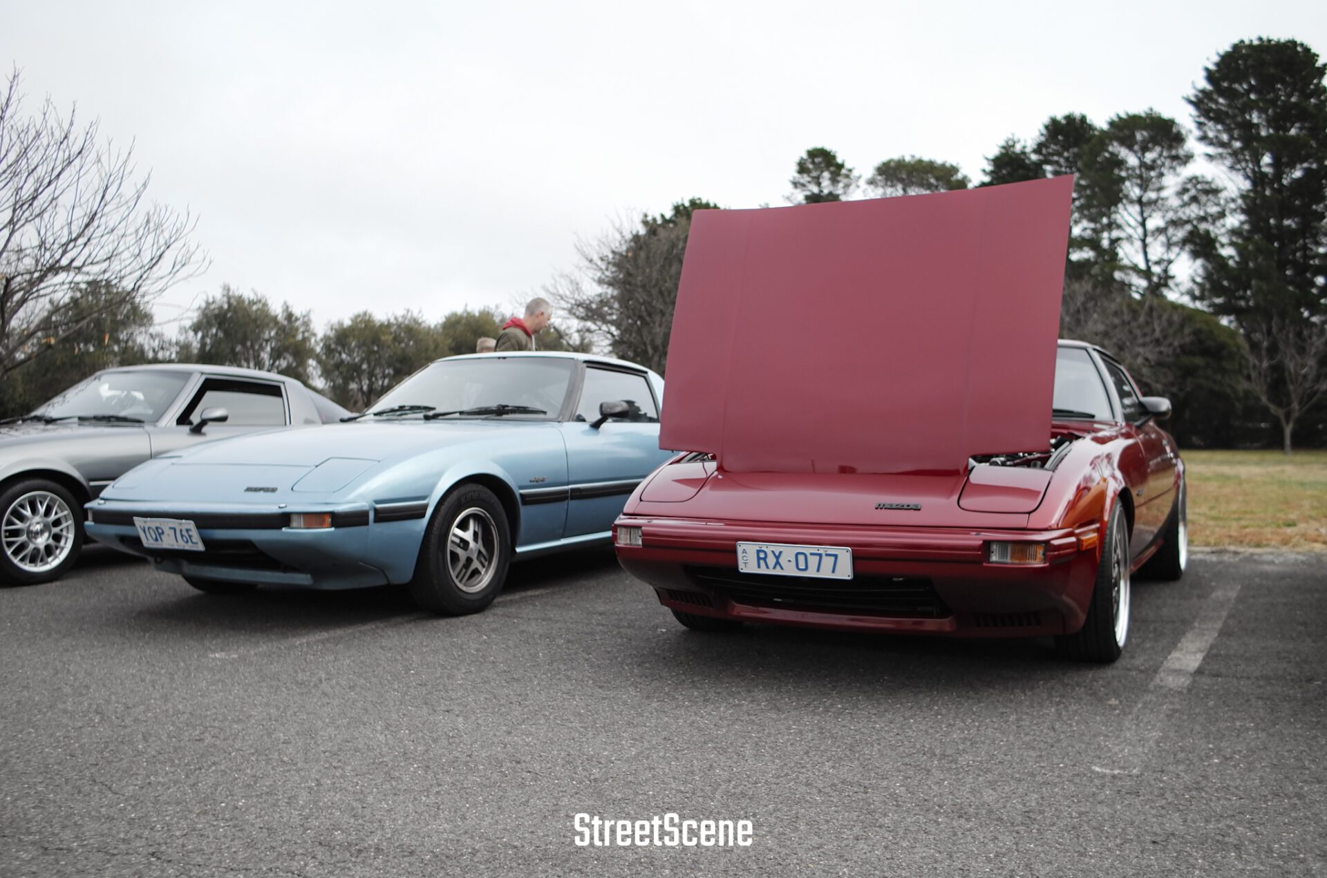 Two vintage Mazda RX-7 cars, one blue and one red with hood open, parked side by side outdoors.