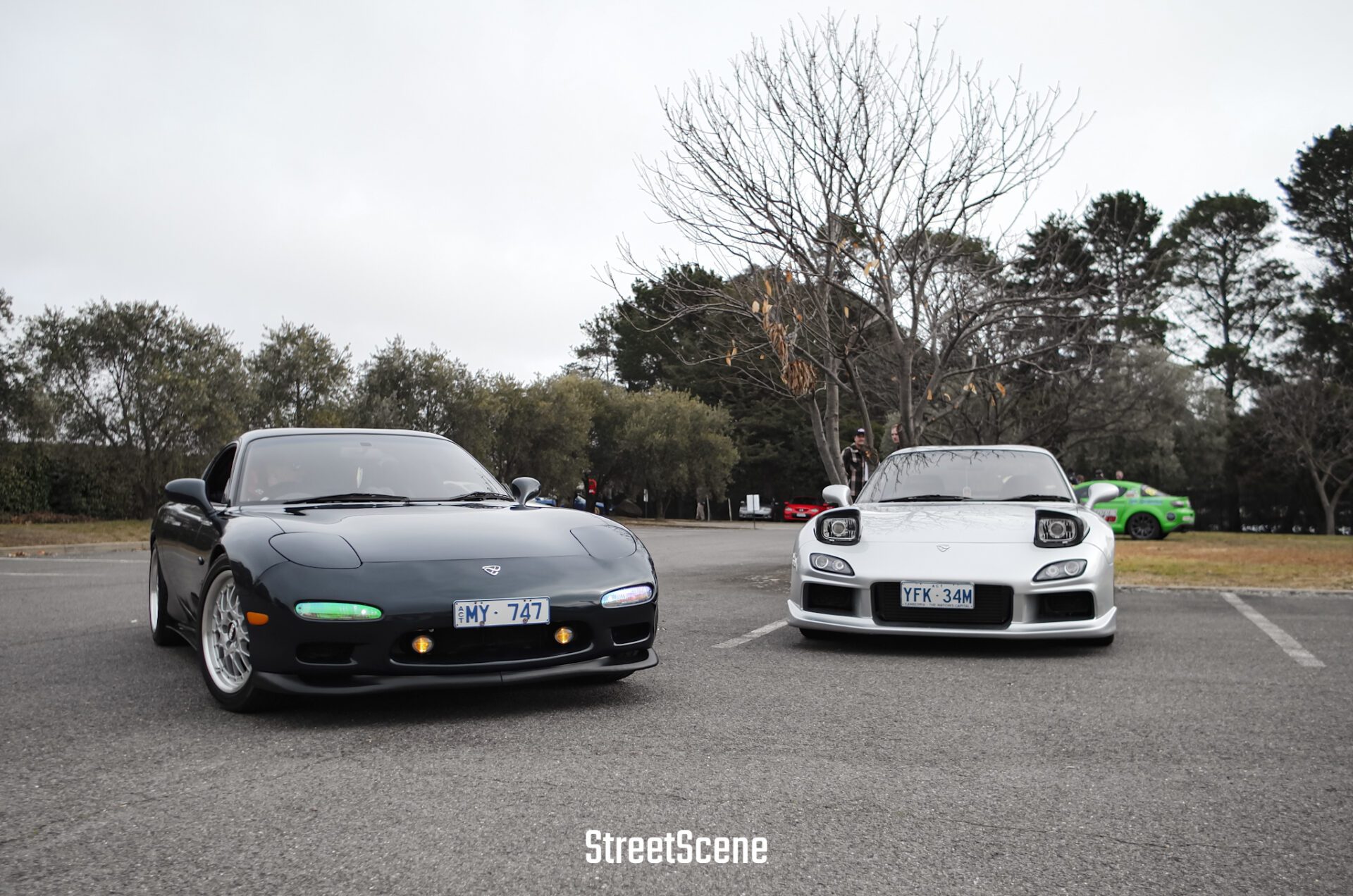 Two sports cars, one black and one white, parked side by side in an outdoor lot with trees.