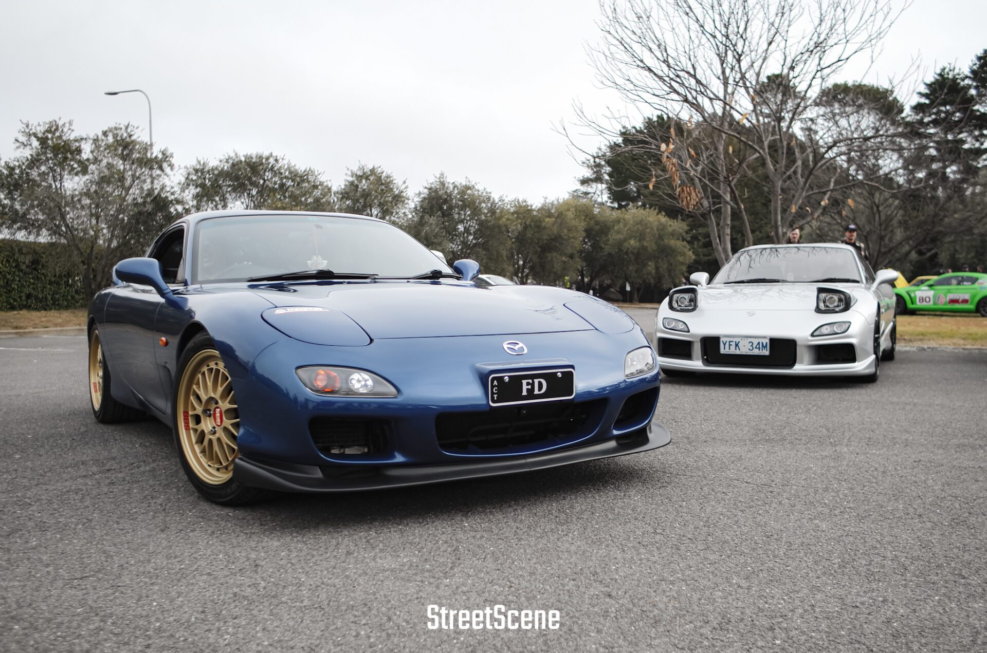 Two Mazda RX-7 sports cars parked outdoors, one blue with gold wheels, one white with pop-up headlights.