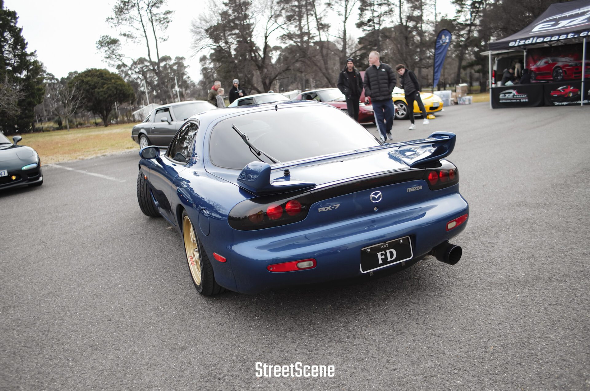 Blue Mazda RX-7 sports car parked with other cars and people in a casual outdoor car meet.
