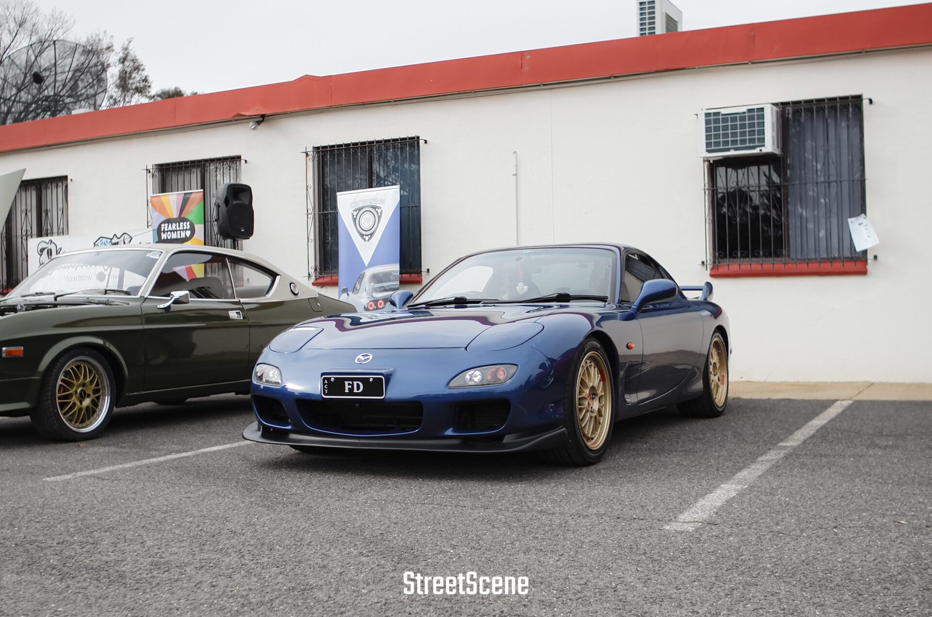 Blue Mazda RX-7 with gold rims parked next to a vintage car in front of a white building.