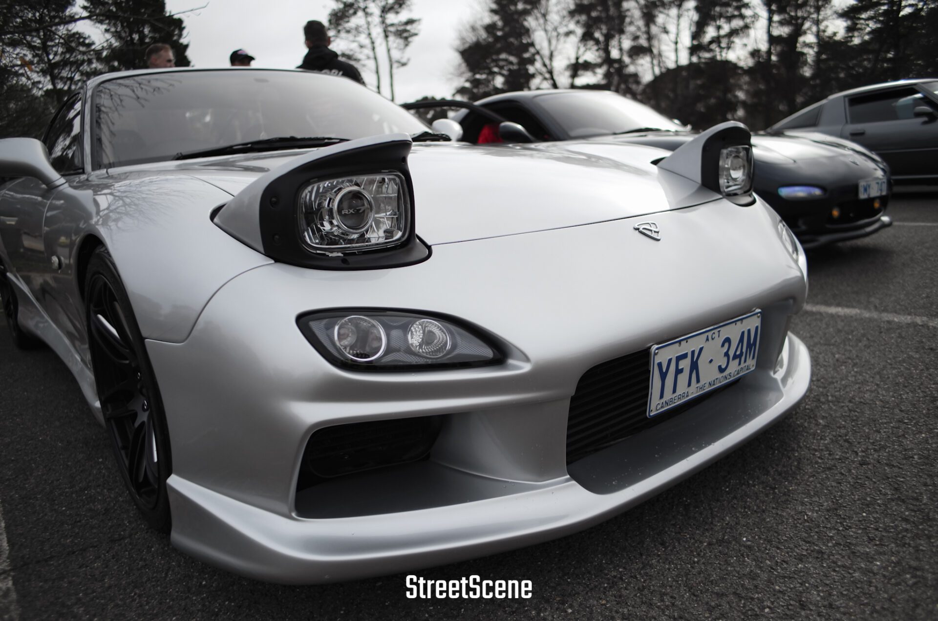 Close-up of a silver Mazda RX-7 with pop-up headlights on a street, with people and other cars in background.