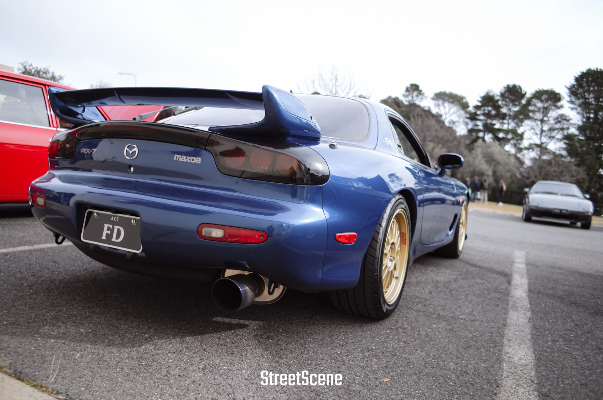 Rear view of a blue Mazda RX-7 with a large spoiler and gold wheels parked outdoors.