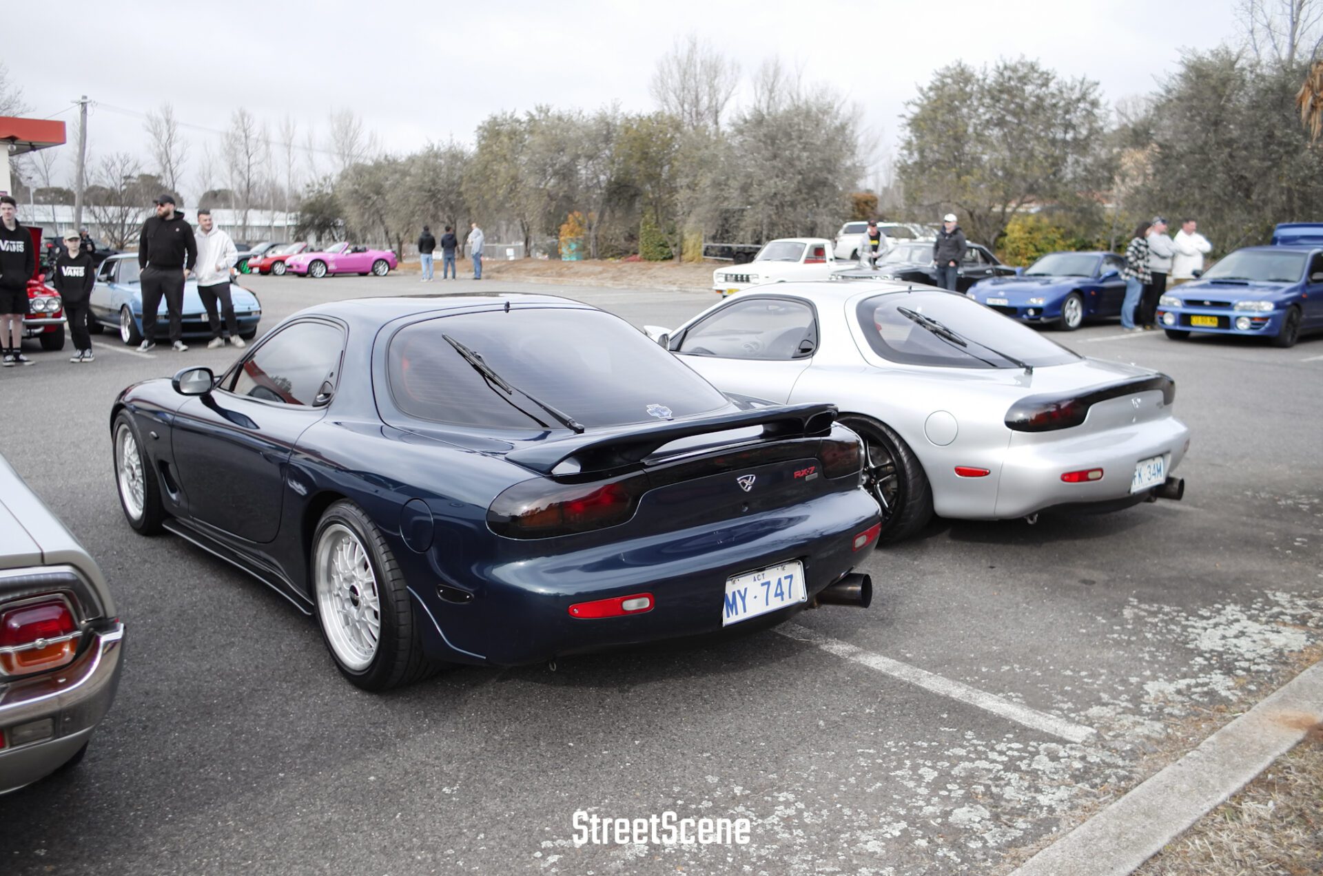 Two Mazda RX-7 cars parked side by side at a car meet with people and other vehicles in the background.