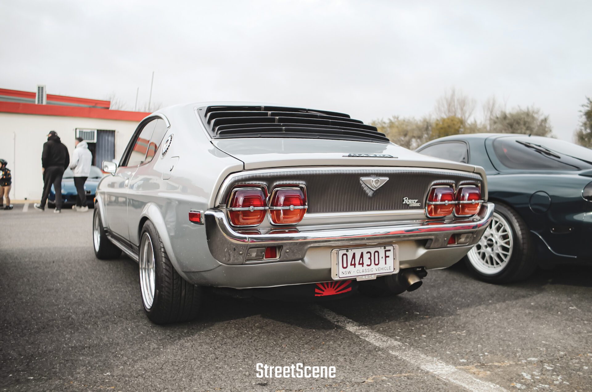 Rear view of a silver classic Mazda RX-3 car with black rear window louvers, parked next to a black sports car.