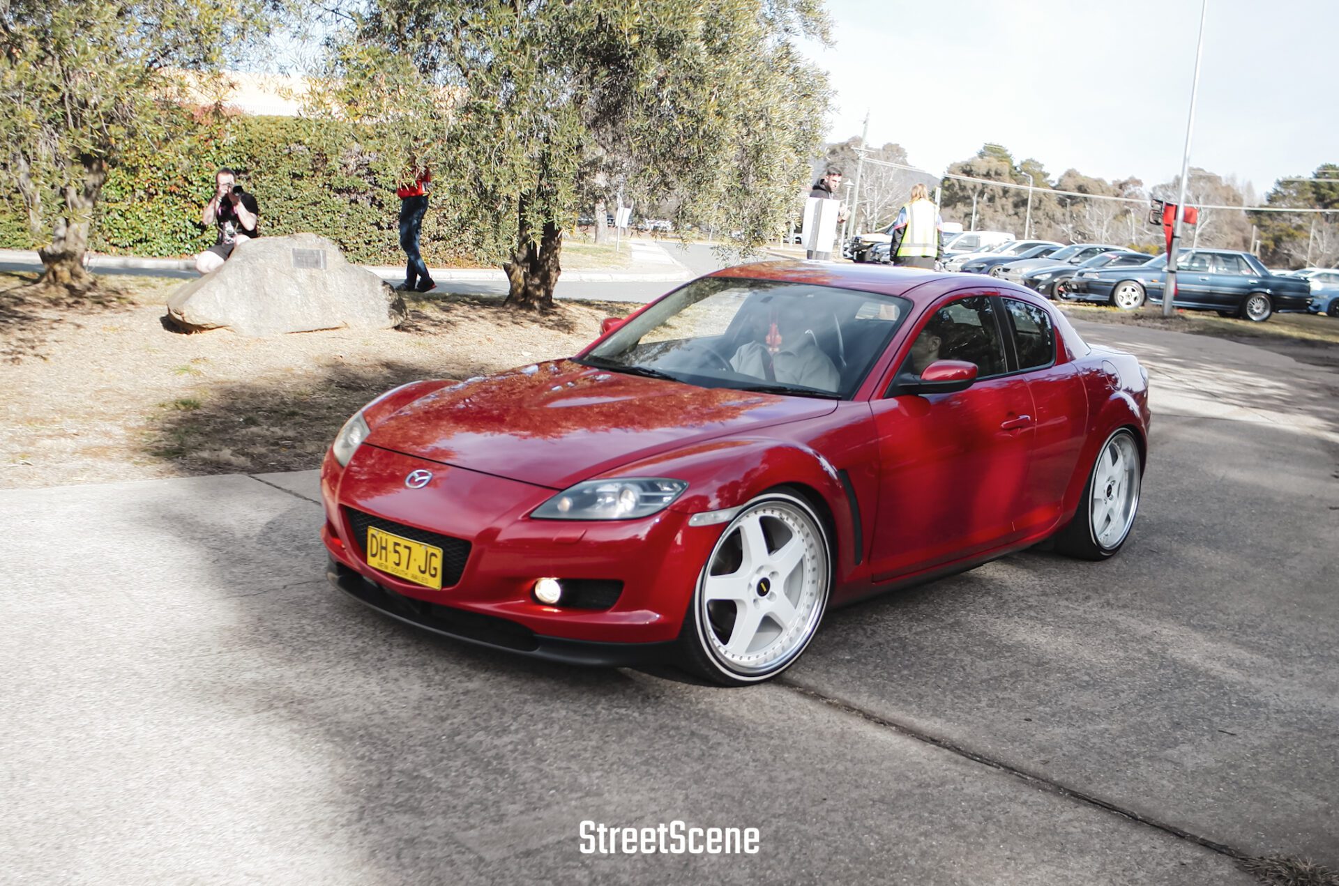 Red Mazda RX-8 with white rims parked on a paved road near trees and onlookers.