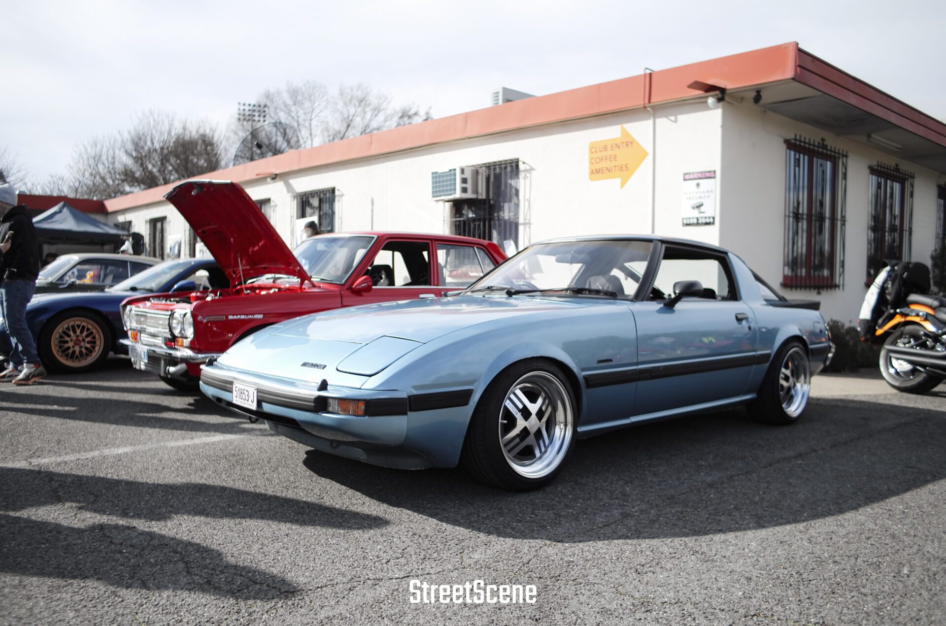 Light blue Mazda RX-7 parked next to a red classic car with its hood open at a car meet.