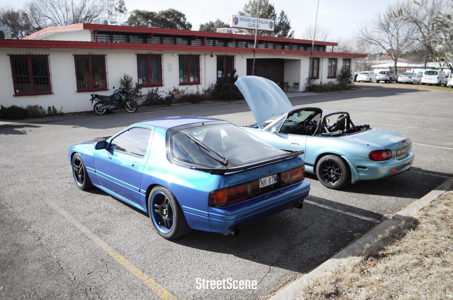 Two blue sports cars parked in a lot near a building with a "Spanish Australian Club" sign.
