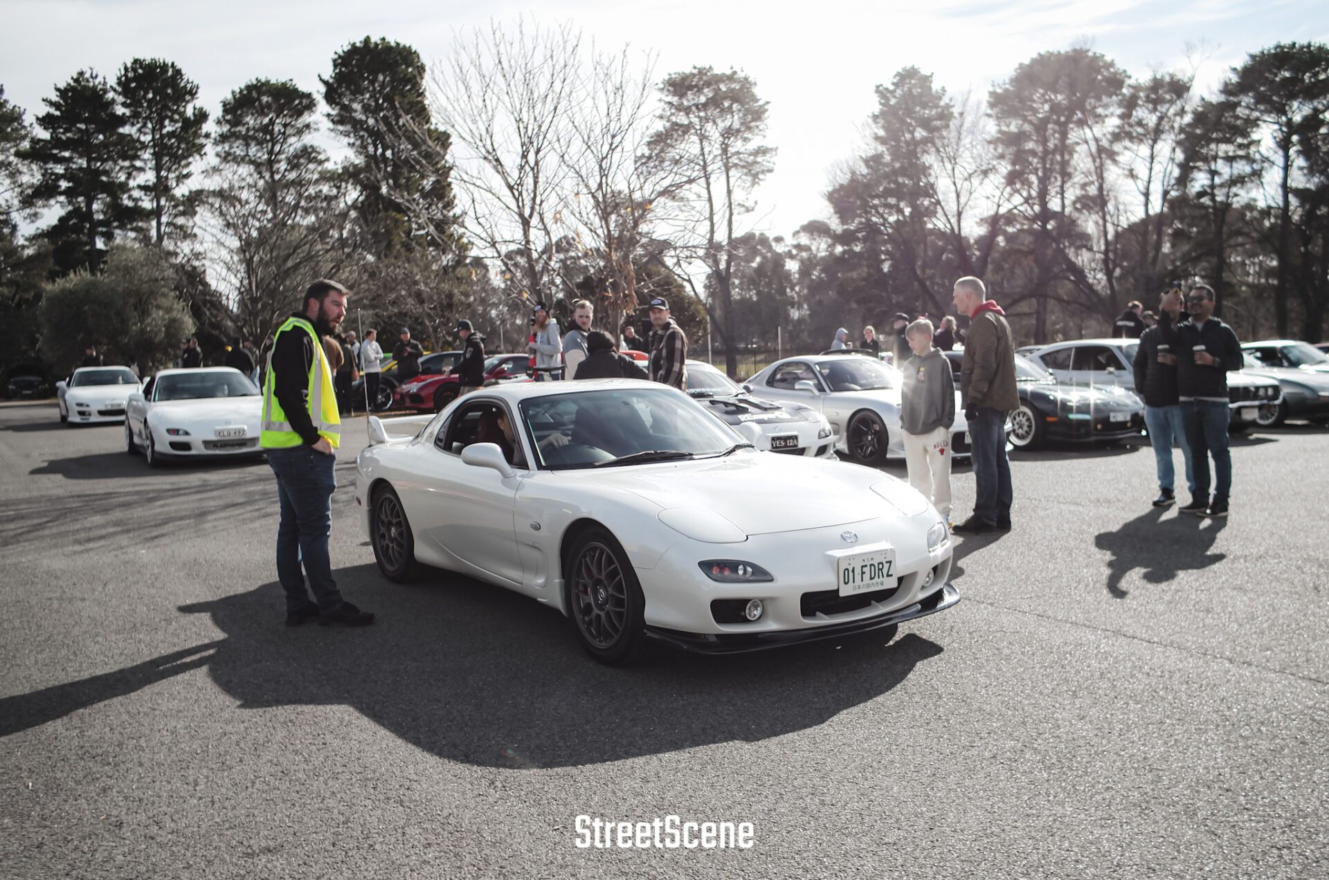 White sports cars parked with people standing nearby at an outdoor car meet under trees.