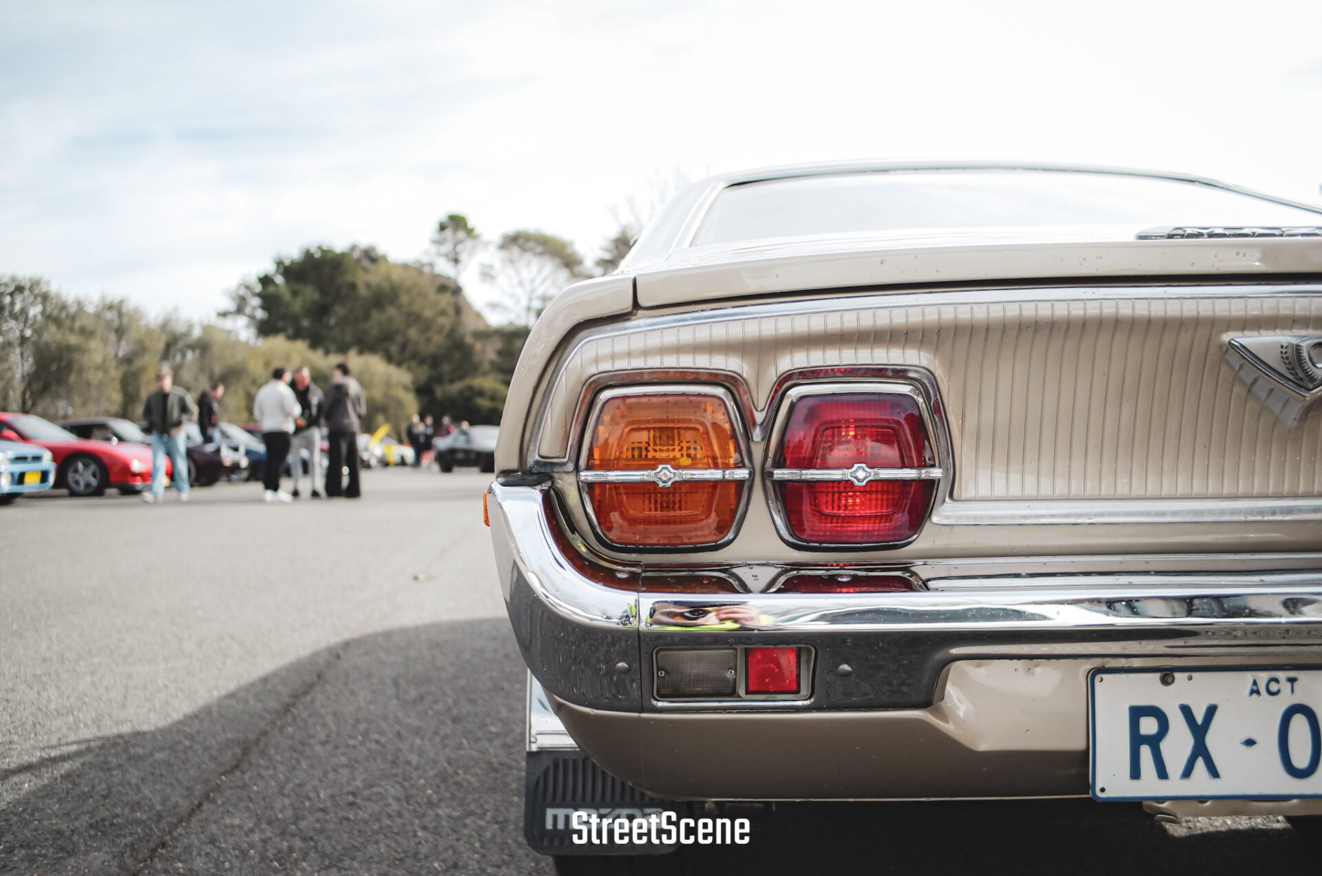 Close-up of a vintage car's rear lights and bumper at an outdoor car meet with people in the background.