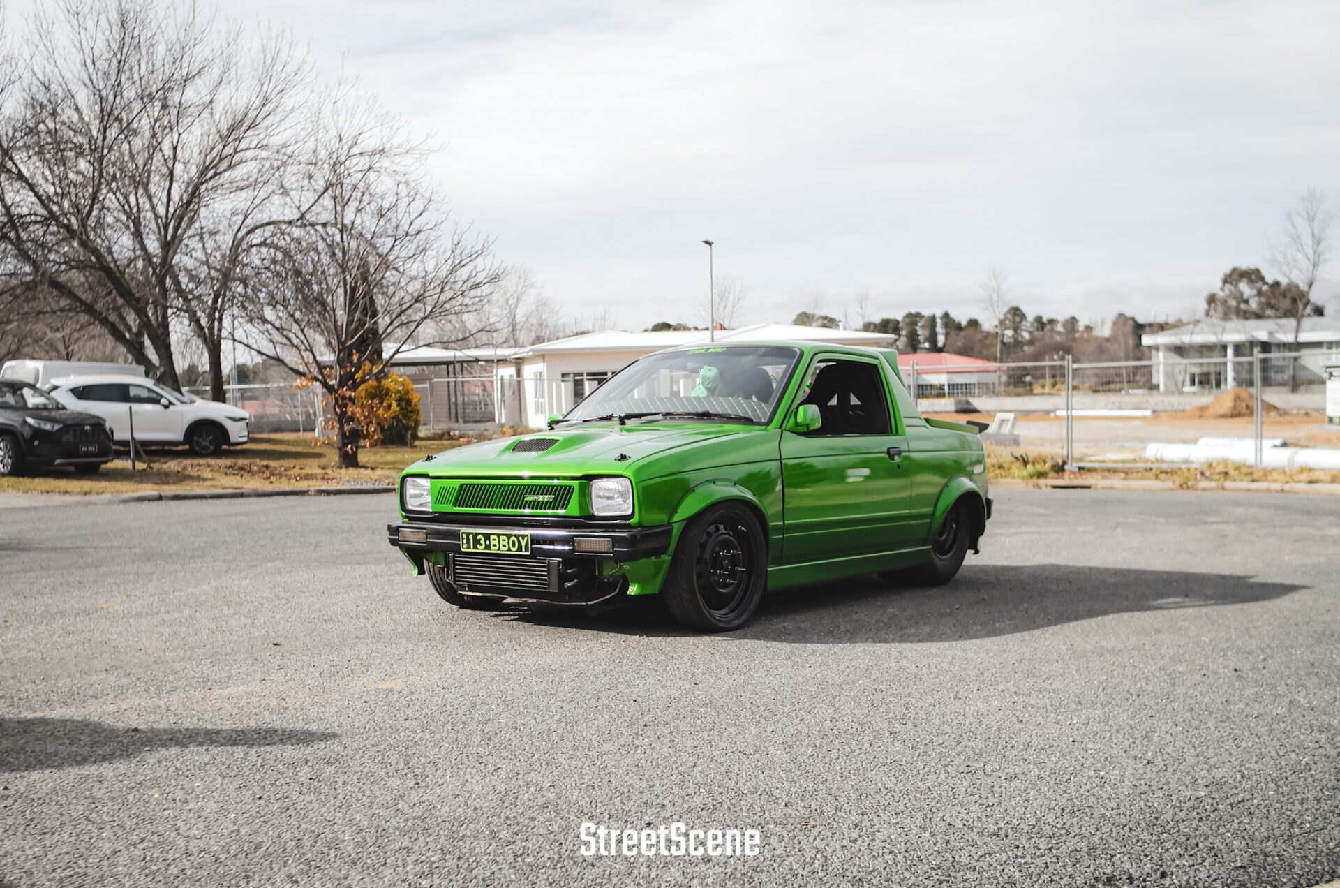 Bright green modified small pickup truck parked on street with bare trees and buildings in background.