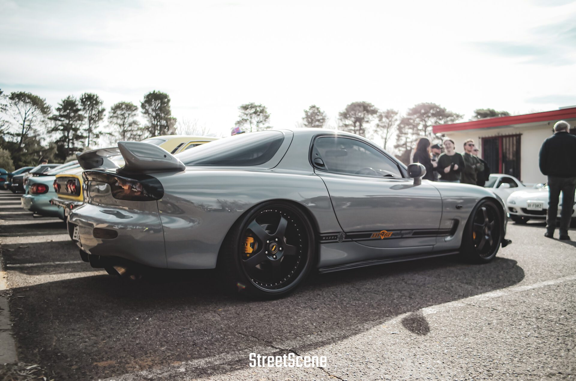Silver Mazda RX-7 with black wheels parked at a car meet with people in the background.
