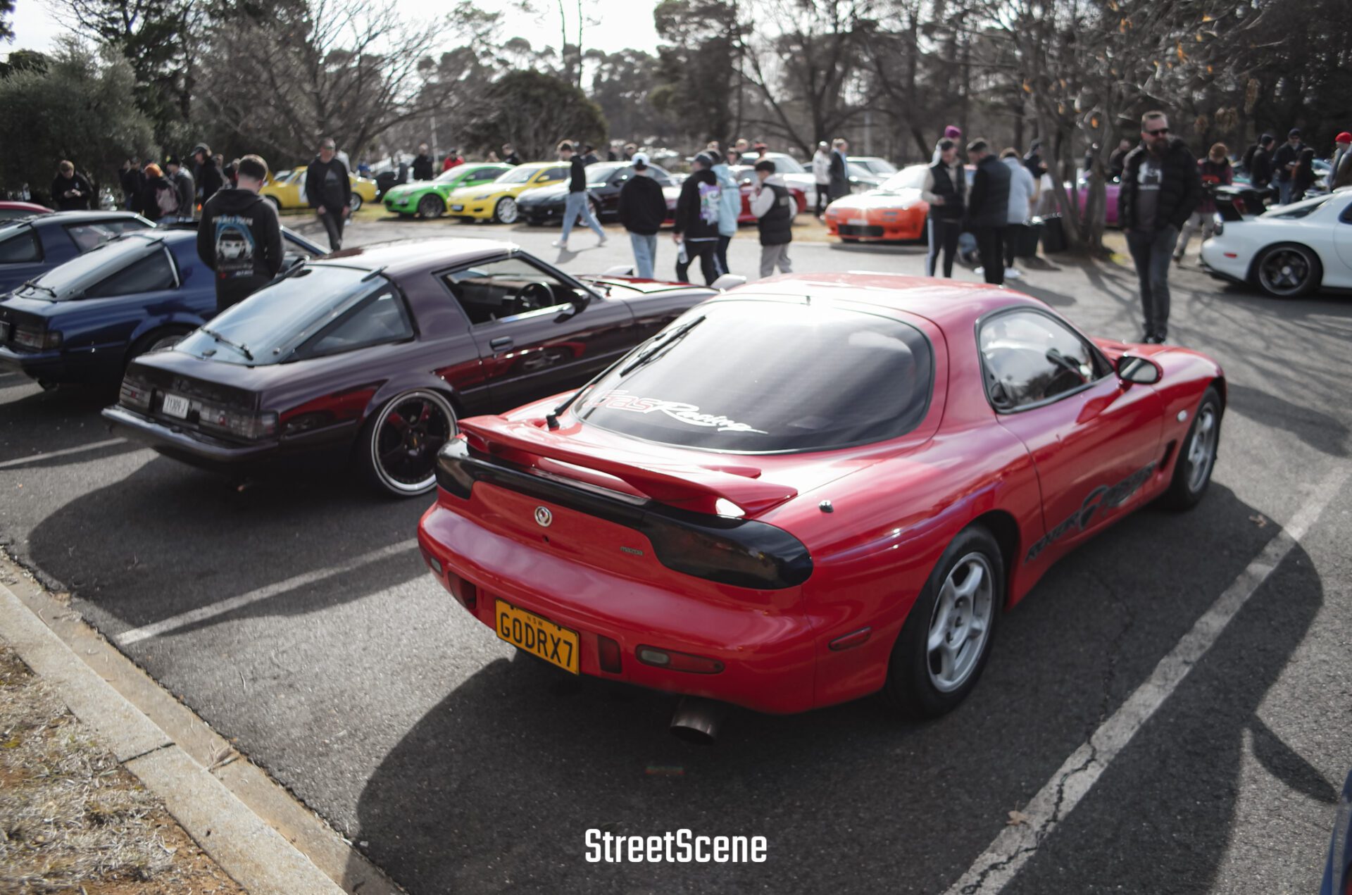 Red Mazda RX-7 and a purple sports car parked at a car meet with people walking around.