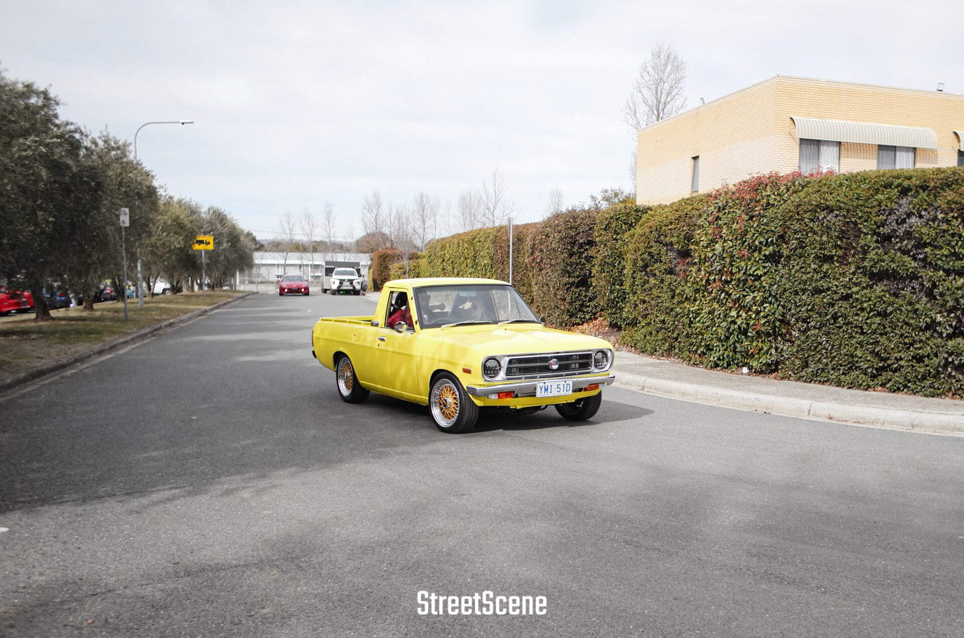Yellow vintage pickup truck driving on an empty street with trees and bushes on the sidewalk.