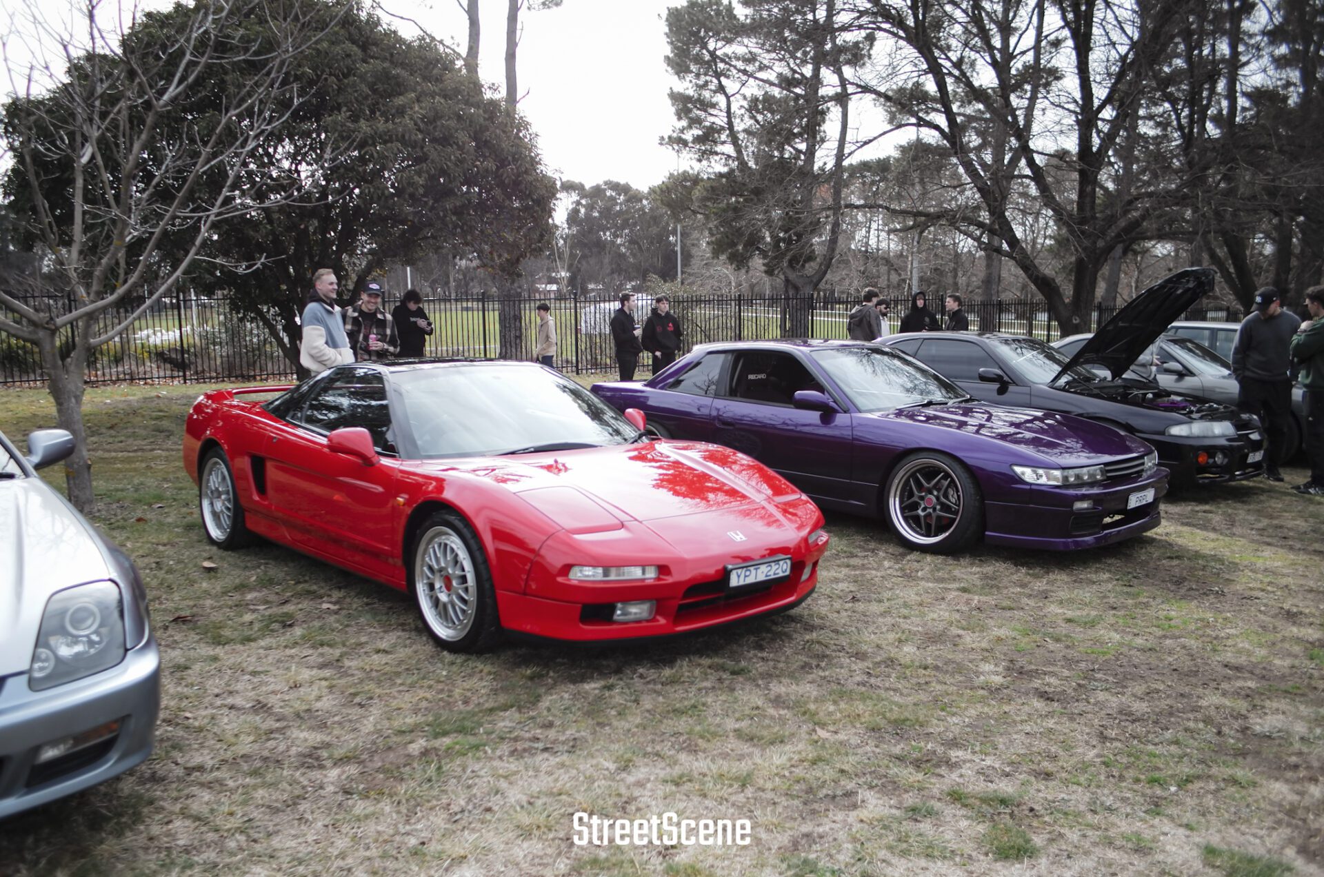 Red Honda NSX and purple Nissan Silvia parked on grass with people in the background at a car meet.