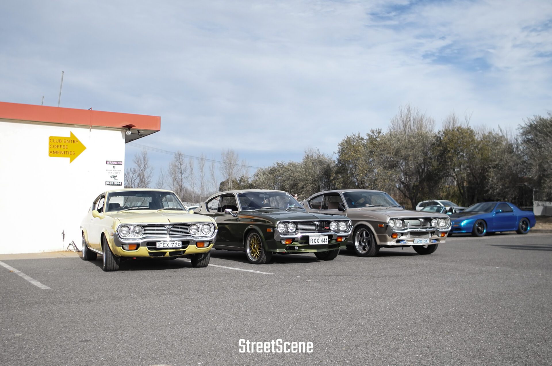 Three vintage cars parked outdoors near a white building with a yellow directional sign.