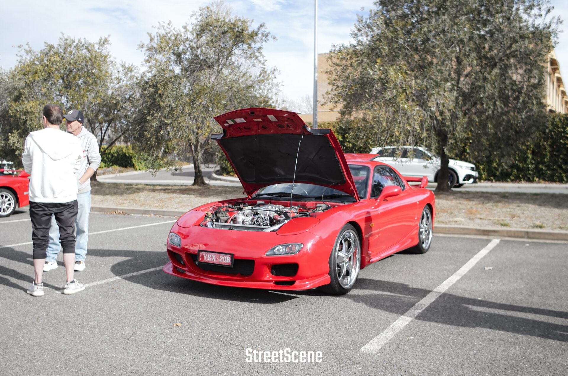 Red sports car with hood open in parking lot, two men standing nearby, trees in background.