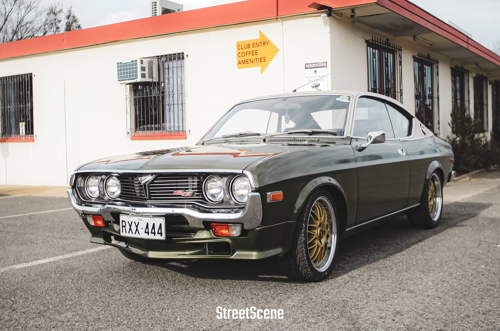 Dark green vintage car with gold rims parked in front of a white building with red trim.